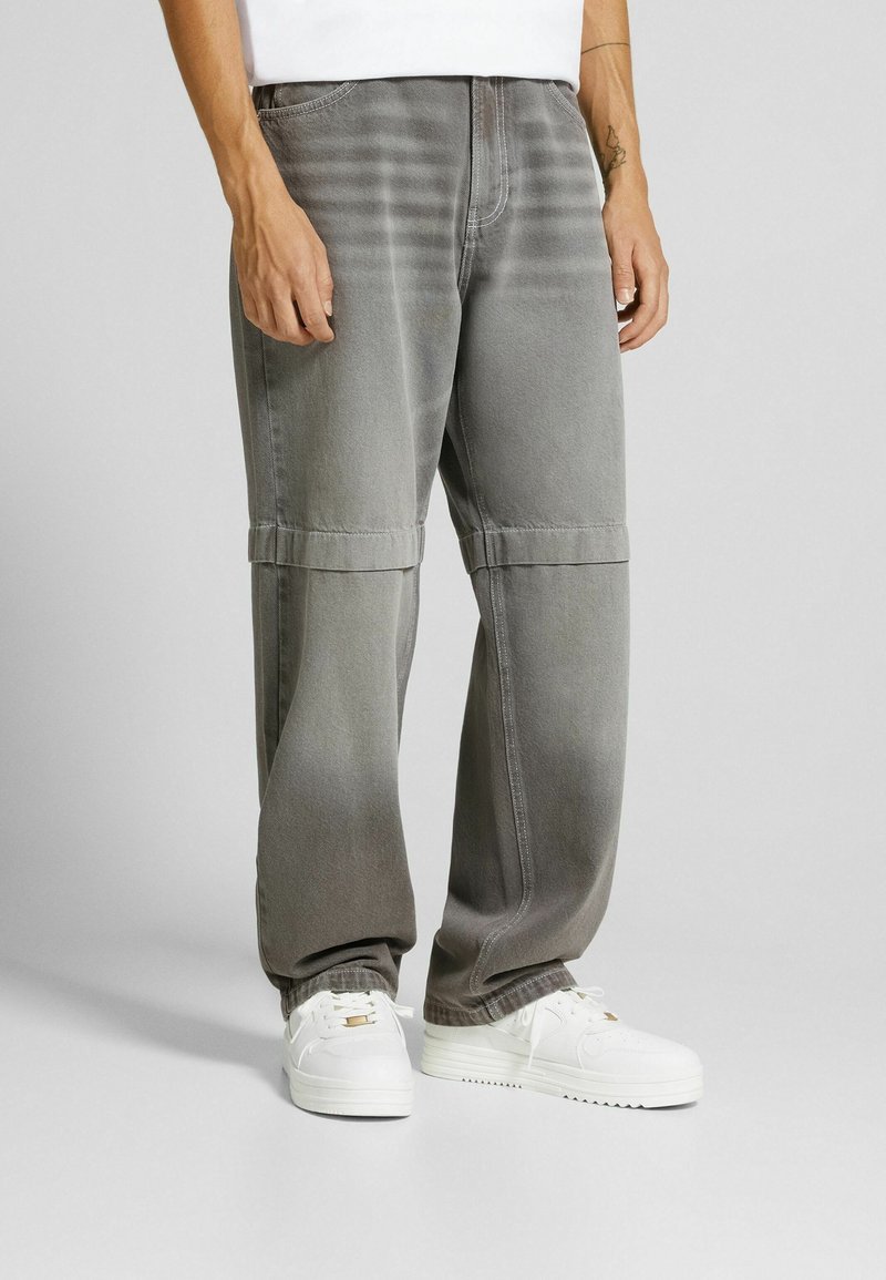 Man som bär lösa grå byxor med sömdetalj ovanför knäna och vita sneakers, stående på en jämn ljusgrå yta.