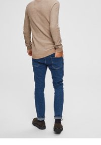 Haut beige en maille à manches longues associé à un jean en denim bleu. Le jean présente un design à cinq poches. La tenue est complétée par des chaussures marron foncé.