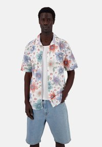 Camicetta a maniche corte con bottoni, su sfondo bianco e grandi motivi floreali colorati. Abbinata a pantaloni corti azzurri.