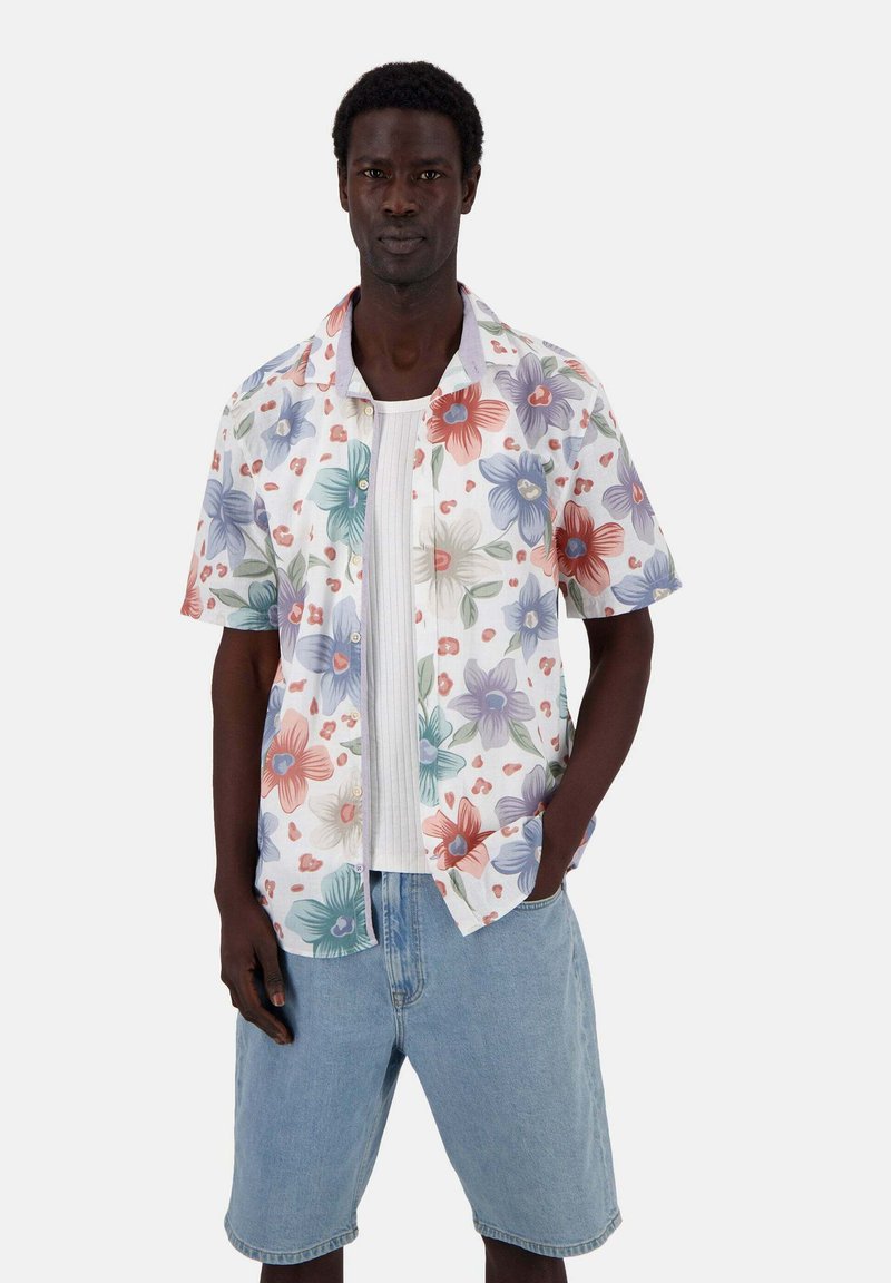 Camicetta a maniche corte con bottoni, su sfondo bianco e grandi motivi floreali colorati. Abbinata a pantaloni corti azzurri.