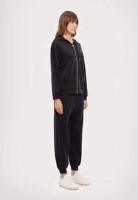 Felpa nera con zip e tasche frontali, dettagli in metallo dorato, abbinata a pantaloni tapered coordinati, indossata con sneakers bianche. Tessuto dalla texture liscia.