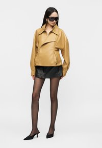 Woman wearing oversized tan leather jacket, black leather mini skirt, sheer black tights, black heeled shoes, and large black sunglasses, standing.