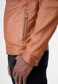 Brun læderjakke med en lynlåslomme på siden, synlige sømme og trykknapdetalje på manchetten. Glat tekstur og tætsiddende design.