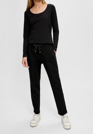 Kvinde iført sort langærmet top og sorte bukser med bindebånd samt hvide sneakers, stående foran en ensfarvet lys baggrund.