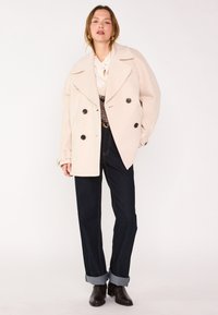 Cappotto di lana doppiopetto color crema con grandi bottoni neri, abbinato a jeans in denim scuro e stivaletti neri, con risvolti arrotolati.