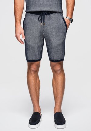 Mann trägt marineblaue und weiß gestreifte Shorts mit Kordelzug, schwarze Slipper mit weißen Sohlen und eine Armbanduhr am linken Handgelenk.