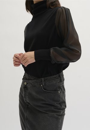 Personne portant un haut noir à manches longues transparentes et un jean en denim gris foncé taille haute, debout devant un fond uni.