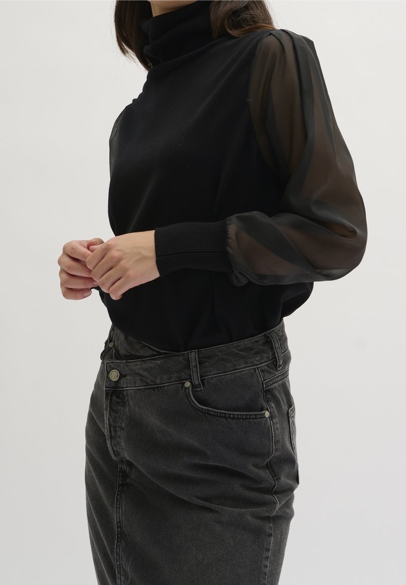 Personne portant un haut noir à manches longues transparentes et un jean en denim gris foncé taille haute, debout devant un fond uni.