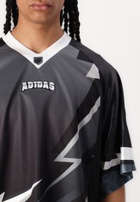 Musta ja harmaa urheilupaita, jossa on V-kaulus, verkkokudos, salaman muotoinen design ja kirjailtu "ADIDAS"-logo rinnassa.