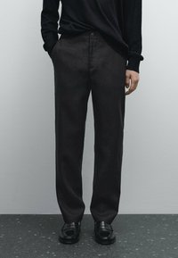 Black linen trousers with a straight fit, featuring side pockets and a button closure. Worn with a black knit top and black loafers.