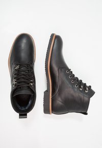 Botas de cuero negro hasta el tobillo con un acabado texturizado, que presentan cordones redondos, ojales de metal, una lengüeta y una suela marrón en contraste.