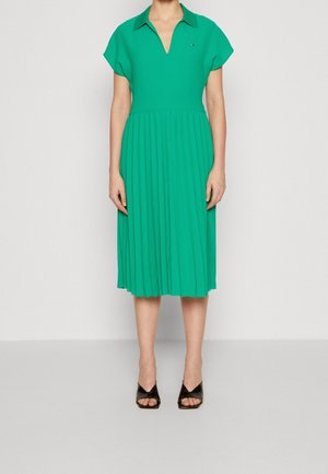 Robe midi plissée verte à manches courtes et décolleté en V. Elle présente un col et un tissu texturé. Chaussures à talons ouvertes noires.