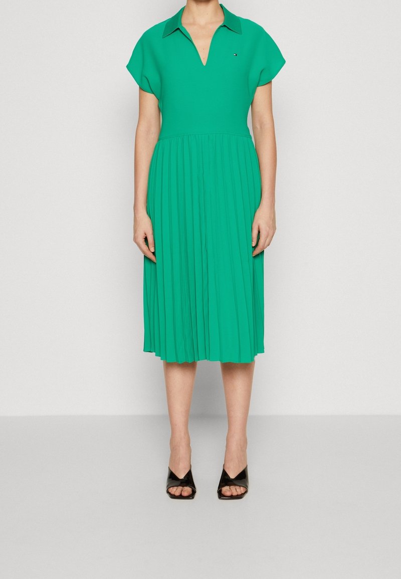 Vihreä pleidattu midi-mekko, jossa on lyhyet hihat ja V-kaula-aukko. Mekossa on kaulus ja teksturoitu kangas. Mustat avonaiset nilkkurit.