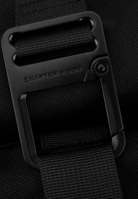 Černý nastavitelný popruh s matně černou kovovou sponou, s vyraženým logem "Kapten & Son" na texturovaném látkovém pozadí.