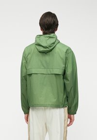 Πράσινο ελαφρύ μπουφάν με κουκούλα, που διαθέτει πίσω τσέπη, ελαστικούς καρπούς και λείο, ματ φινίρισμα.