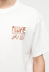 Valge T-särk oranži tikandiga "NIKE MUSIC ENERGY" ja nelja muusikamärgi sümboliga vasakul rinnal.