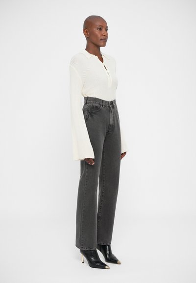 Persona vistiendo una blusa de canalé crema de manga larga con puños acampanados, jeans oscuros de cintura alta y botas de tacón negras de punta. 