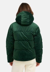 Groene fluwelen pufferjack met een capuchon, met een doorgestikt ontwerp en ribbelstructuur. Gedragen over lichtgekleurde broeken.