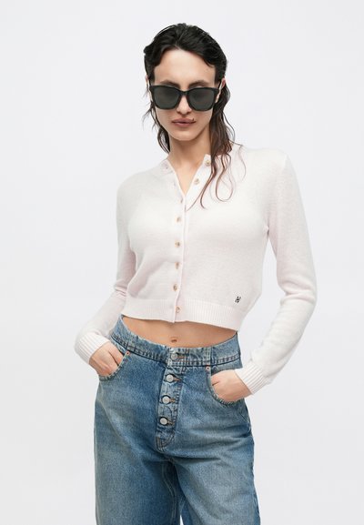 Világos rózsaszín rövidített kardigán gombos elülső résszel és bordázott mandzsettákkal, magas derekú kék farmernadrággal párosítva, amely gombos záródású.