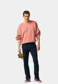 Sweat-shirt surdimensionné rose, pantalon bleu marine et baskets violettes. Modèle tenant un petit bouquet de fleurs. Fond simple.
