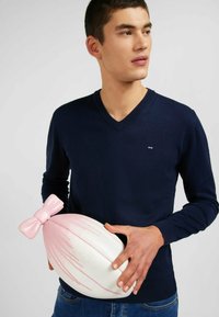 Pull-over v-ni de couleur marine, en tissu tricoté, au design ajusté. Le mannequin tient un grand objet ovale blanc avec un nœud rose et des traits, ressemblant à un élément décoratif.