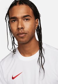 Maglietta sportiva bianca con scollatura rotonda e maniche corte; presenta il logo rosso di Nike sul lato sinistro del petto. Capelli acconciati in lunghe trecce.