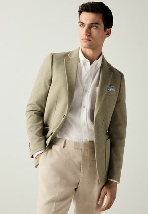 Beige blazer with textured fabric and two buttons, over a white linen shirt. Trousers are light-colored. Pocket square adds color detail.