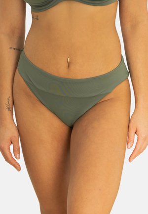 Frau trägt olivgrünen Bikiniober- und unterteil, zeigt die Körpermitte und die Oberschenkel mit sichtbaren Tätowierungen an den Armen und einem Bauchnabelpiercing.