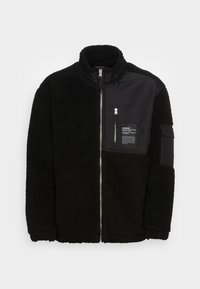Giacca in pile nera con esterno testurizzato, dotata di zip frontale, colletto alto e un pannello in nylon nero con tasca a zip e etichetta.