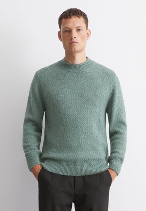 Grüner strukturierter Strickpullover mit Rundhalsausschnitt und gerippten Bündchen, kombiniert mit dunklen Streifenhosen.