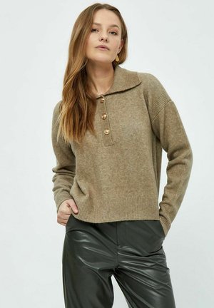 Beige stickad tröja med bred krage, fyra gyllene knappar och ribbad nederkant. Parat med svarta, glänsande, högmidjade byxor.