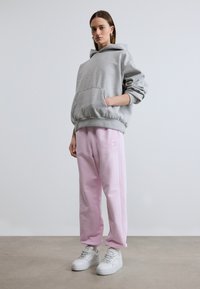 Γυναίκα που φοράει γκρι φαρδύ φούτερ με κουκούλα, ροζ ανοιχτό φόρμα και λευκά αθλητικά παπούτσια, στέκεται σε γκρι πάτωμα μπροστά σε άσπρο τοίχο.