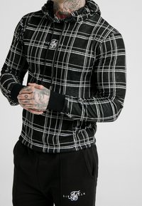 Black and white plaid hoodie with a drawstring, featuring a logo in the center and ribbed cuffs. Paired with black pants displaying a logo.