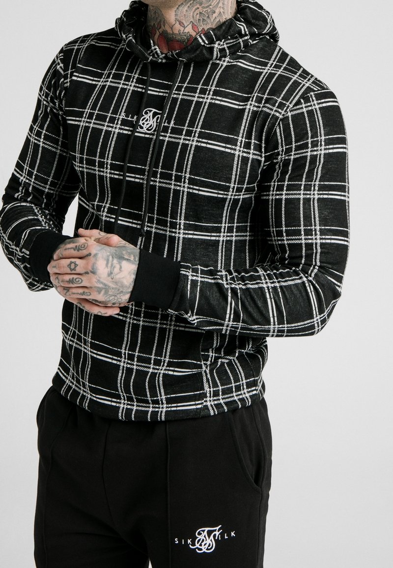 Black and white plaid hoodie with a drawstring, featuring a logo in the center and ribbed cuffs. Paired with black pants displaying a logo.