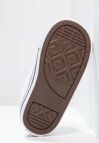 Biała podeszwa sneakerów z brązowym bieżnikiem, z geometrycznymi wzorami i kształtami gwiazd, nachylona względem białej powierzchni.