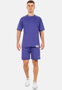 T-shirt viola a maniche corte e pantaloni corti abbinati realizzati in tessuto misto cotone. Presenta un collo a giro e dettagli con logo vicino all'orlo. Sneakers bianche.