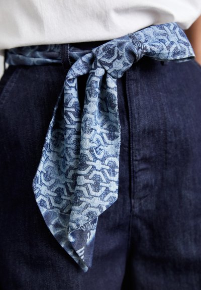 Denim josta ar jostu rozā un gaiši zilo rakstu, sasieta priekšā, kopā ar tumši ziliem augstās jostasvietas biksēm.
