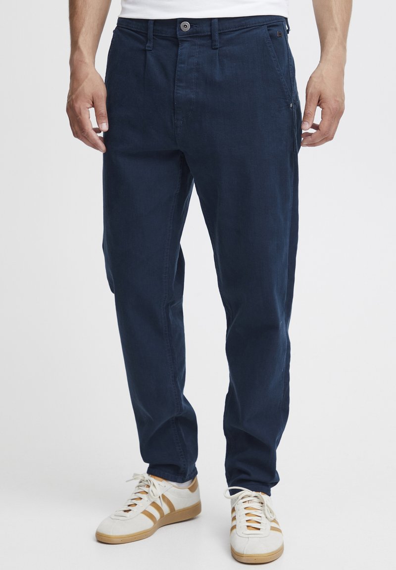 Tumed puuvillased püksid kitsas lõikes, millel on lukuga tasku, nööpidega sulgemine ja küljetaskud, koos valgete tennisersustega.