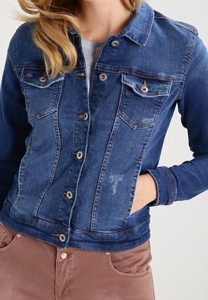 Femme portant une veste en denim bleu avec fermeture à boutons, poches poitrine, une main dans la poche latérale, associée à un pantalon marron.