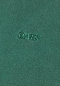 Tessuto verde con una superficie testurizzata che presenta un logo ricamato "Levi's". L'immagine si concentra sul materiale e sui dettagli del logo.