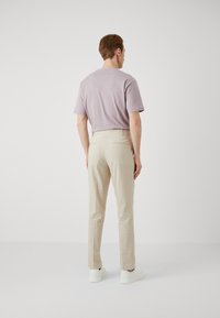 Achteraanzicht van een persoon in een lavendel kleurig t-shirt met korte mouwen en beige op maat gemaakte broek, gecombineerd met witte sneakers, neutrale achtergrond.