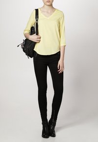 Blouse jaune à manches longues avec un décolleté en V, portée avec un jean skinny noir et des bottines. Un sac à dos noir est porté sur une épaule.