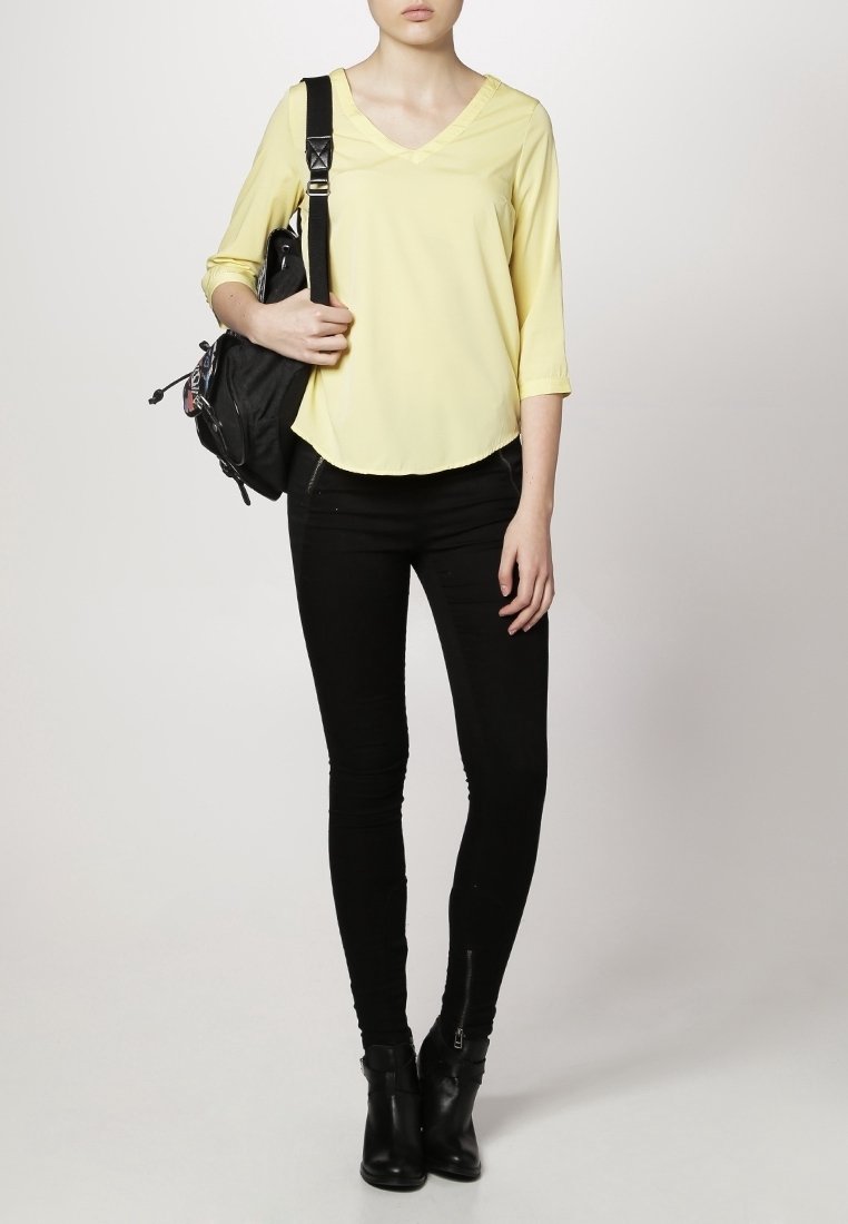 Blouse jaune à manches longues avec un décolleté en V, portée avec un jean skinny noir et des bottines. Un sac à dos noir est porté sur une épaule.