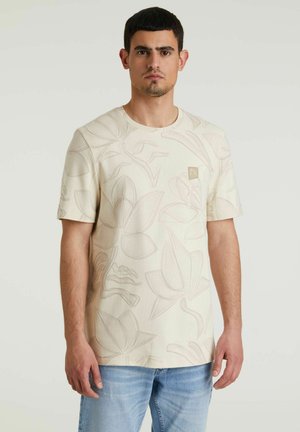 Beige T-Shirt mit floralem Reliefmuster, kurzen Ärmeln, Rundhalsausschnitt und kleinem Logo-Patch auf der Brust. Getragen mit hellblauen Jeans.