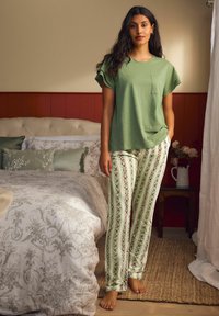 Zaļš kokvilnas T-krekls ar īsām piedurknēm un krūšu kabatu, kopā ar rakstainām pidžamas biksēm ar vertikālām svītrām un ziedu motīviem.