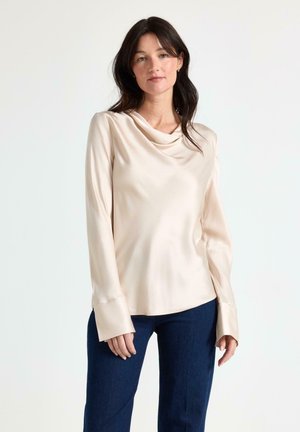 Kvinde med mørkt hår iført en langærmet cremefarvet satinbluse og mørkeblå jeans, stående mod en ensfarvet lys baggrund.