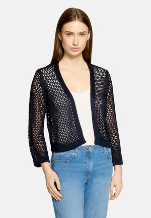 Kvinde iført en sort åbenstrikket cardigan over en hvid top og blå jeans, stående mod en lys grå baggrund.