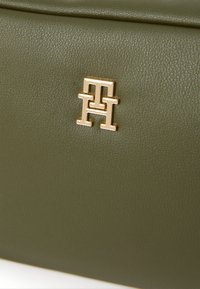 Olive green textured leather bag featuring a prominent gold-tone logo embossed on the surface. Compact and rectangular shape.