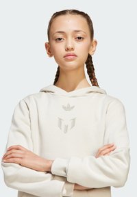 Sweatshirt à capuche crème avec une texture douce, présentant un logo en relief bien en vue et un design minimaliste, porté par un modèle les bras croisés.