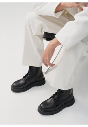 Stivali neri in pelle con lacci, punta arrotondata, suola spessa in gomma e design minimalista. Indossati con pantaloni bianchi su uno sfondo chiaro.